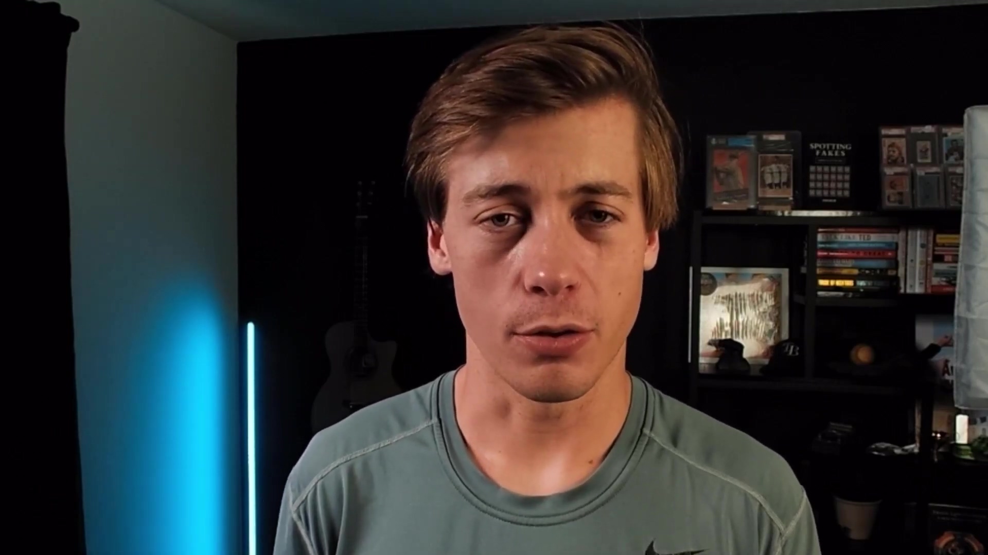 The image shows a young man, likely the video presenter, looking directly at the camera. He has short, light brown hair and is wearing a teal-colored t-shirt with a black Nike logo on the right chest area. His expression is neutral, and he appears to be speaking. The background is dark, with a black wall behind him. To his left, a light-colored acoustic guitar is partially visible. To his right, a black shelf unit holds various items, including what appear to be framed trading cards or collectibles, and some books. A blue light emanates from the left side of the frame, casting a subtle glow.