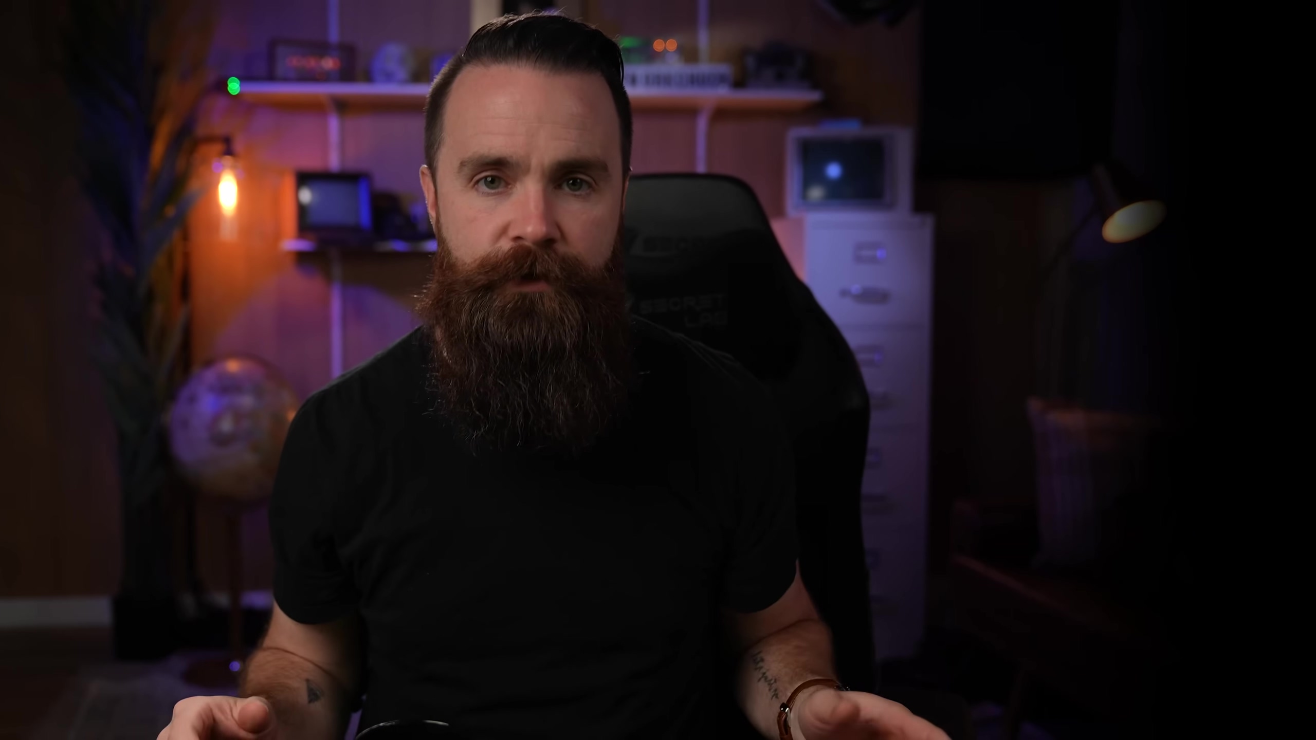 A man with a full, reddish-brown beard and dark hair is centered in the frame, looking directly at the camera. He is wearing a black t-shirt. The background is dimly lit with warm, ambient lighting, featuring shelves with various objects, including a globe on the left, a lamp with an exposed bulb, and what appears to be a server rack or computer equipment on the right. A black gaming chair with 'SECRET LAB' visible on the headrest is behind him. The overall mood is serious and engaging.