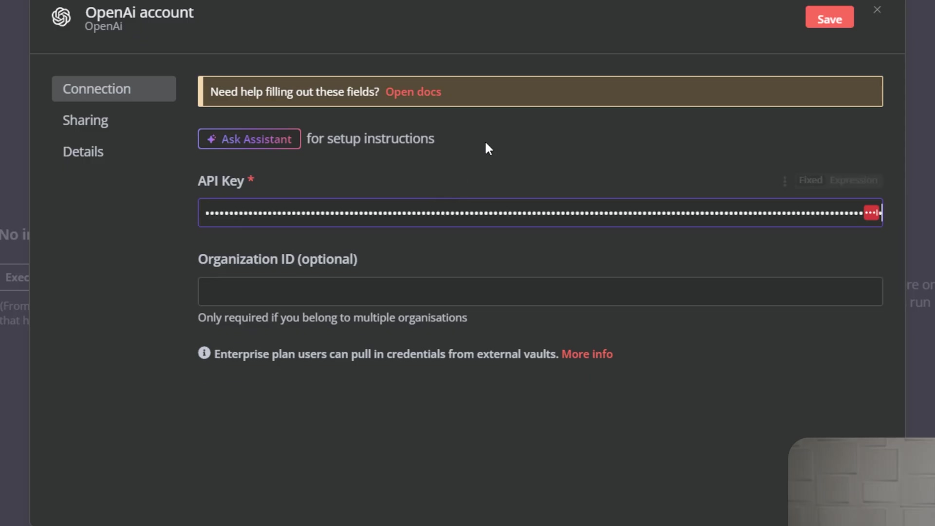 The image shows a dark-themed interface for an 'OpenAI account' connection. The left sidebar has navigation options: 'Connection', 'Sharing', and 'Details', with 'Connection' currently selected. The main content area features a banner stating 'Need help filling out these fields? Open docs'. Below this, there's a button labeled 'Ask Assistant' for setup instructions. The primary input fields are 'API Key*' (obscured with dots, indicating a sensitive value) and 'Organization ID (optional)'. A note below the Organization ID field clarifies 'Only required if you belong to multiple organisations'. Further down, an informational message states 'Enterprise plan users can pull in credentials from external vaults. More info'. A 'Save' button is in the top right, and an 'x' button for closing is next to it. A man is partially visible in the bottom right corner.