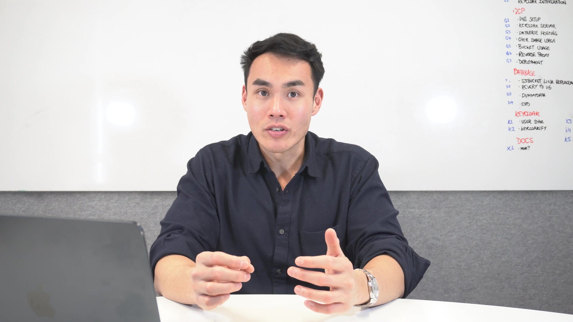 A man with short dark hair and a dark blue collared shirt is seated at a desk, looking directly at the camera and speaking. He has his hands clasped together in front of him. To his left, a silver laptop is partially visible. Behind him is a white board with handwritten notes in blue and red markers. The notes on the whiteboard are partially visible and include headings like 'KEYCLOAK INTEGRATION', 'DCP', 'DATABASE', and 'DOCS'. Under 'DCP', items like 'G1 - REVERSE SERVER', 'G2 - DATABASE HOSTING', 'G3 - DOCKER IMAGE', 'G4 - BUCKET', 'G5 - REVERSE PROXY', 'G6 - DEPLOYMENT' are listed. Under 'DATABASE', items like 'Z1 - SUBGRAPH', 'Z2 - REDIS', 'Z3 - DYNAMODB', 'Z4 - EXD' are visible. Under 'KEYCLOAK', 'K1 - USER SINK', 'K2 - KEYCLOAKIFY' are listed. Under 'DOCS', 'X1 - NNN?' is visible. The lighting is bright, highlighting the man and the whiteboard.