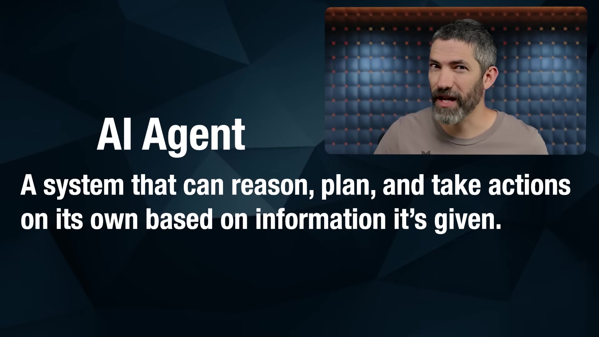 The image displays a definition of an 'AI Agent' on a dark blue background with geometric patterns. The text 'AI Agent' is prominently displayed in large white font at the top left. Below it, in a smaller white font, is the definition: 'A system that can reason, plan, and take actions on its own based on information it's given.' In the upper right corner, a speaker with a beard and short hair is visible, looking directly at the camera. He is wearing a light brown t-shirt and is positioned in front of a dark blue padded wall.