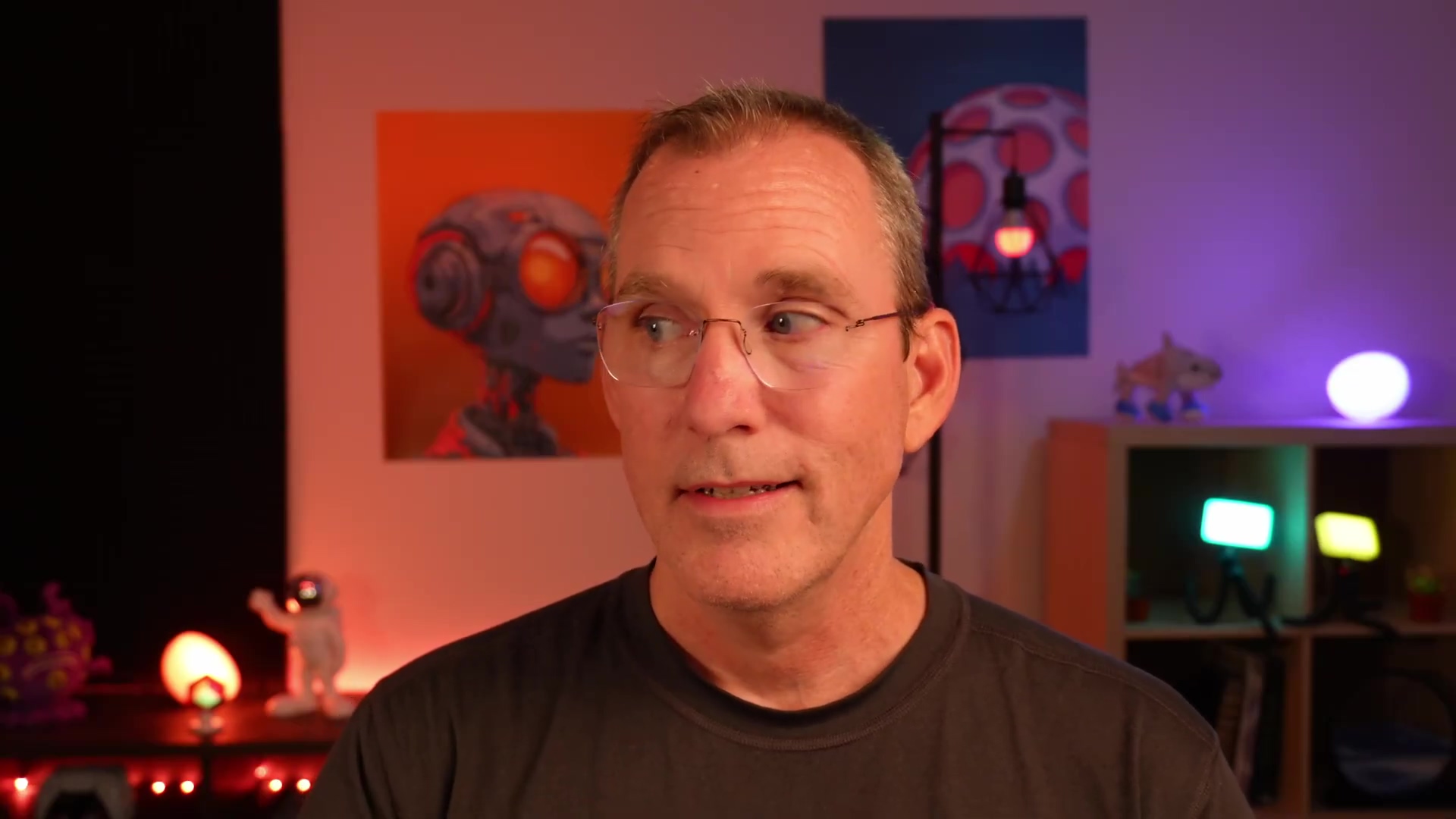 The image shows a man with short graying hair and glasses looking slightly to the right. He is wearing a dark gray t-shirt. The background includes an orange-toned picture of a robot-like figure, a colorful mushroom-shaped lamp, and a small white figure on a shelf. The lighting is warm, with a mix of orange and purple hues.