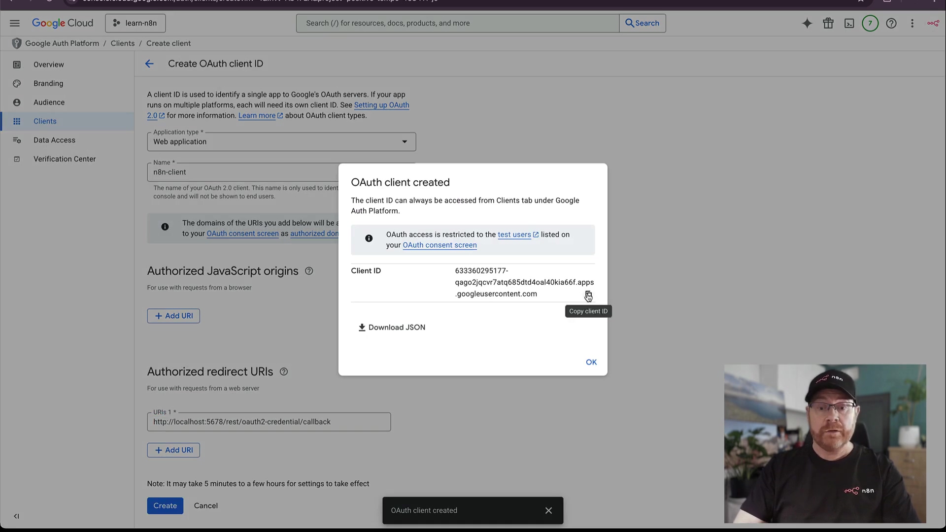 The image displays a Google Cloud Platform interface for creating an OAuth client ID. The main section is titled "Create OAuth client ID." Under "Application type," "Web application" is selected. The "Name" field shows "n8n-client." Below this, there's a section for "Authorized JavaScript origins" and "Authorized redirect URIs," with one URI listed as "http://localhost:5678/rest/oauth2-credential/callback." A pop-up window in the center is titled "OAuth client created" and displays a "Client ID" which is a long alphanumeric string, and options to "Download JSON" or "Copy client ID." It also notes that "OAuth access is restricted to the test users listed on your OAuth consent screen." The bottom left of the screen has "Create" and "Cancel" buttons. A small black notification bar at the bottom reads "OAuth client created." A man with glasses and a black t-shirt with a logo is visible in the bottom right corner of the screen.