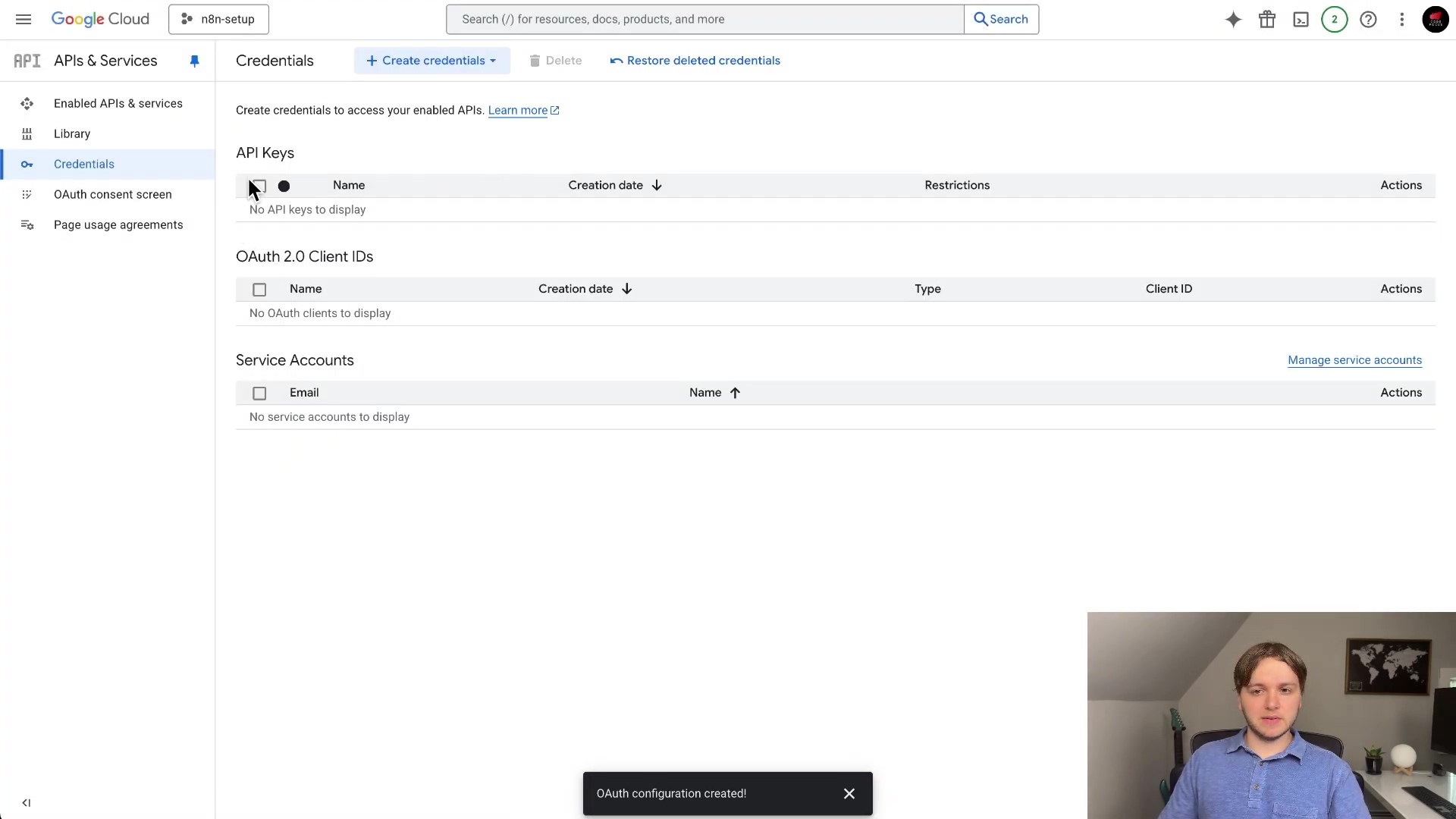 The image shows the 'Credentials' section within the Google Cloud Platform interface. The main content area lists sections for 'API Keys', 'OAuth 2.0 Client IDs', and 'Service Accounts'. Under 'API Keys', it states 'No API keys to display'. Similarly, under 'OAuth 2.0 Client IDs', it says 'No OAuth clients to display'. Under 'Service Accounts', it says 'No service accounts to display'. At the top, there are buttons for '+ Create credentials', 'Delete', and 'Restore deleted credentials'. A notification at the bottom of the screen reads 'OAuth configuration created!'. The left sidebar shows 'API APIs & Services' expanded with 'Library', 'Credentials', and 'OAuth consent screen' as sub-options. A person is visible in the bottom right corner of the screen.