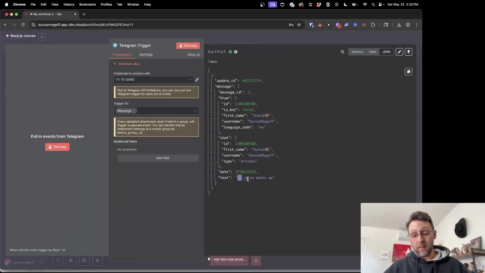 The image displays a split screen of a desktop interface, likely a web browser, showing an n8n workflow on the left and a JSON output on the right. The n8n interface has a 'Telegram Trigger' node selected, with 'Parameters' and 'Settings' tabs visible. Below the node, there's a 'Webhook URLs' section and 'Credential to connect with' set to 'YT Tg demo'. A message below states 'Due to Telegram API limitations, you can use just one Telegram Trigger for each bot at a time.' The 'Trigger On' is set to 'Message'. The right side shows a JSON output with details of a received Telegram message, including 'update_id', 'message_id', 'from' user details ('first_name': 'DuncanG', 'username': 'DuncanRogoff', 'language_code': 'en'), 'chat' details ('first_name': 'DuncanG', 'username': 'DuncanRogoff', 'type': 'private'), 'date', and 'text': 'yo yo whats up'. A person with glasses is visible at the bottom right, looking at the screen.