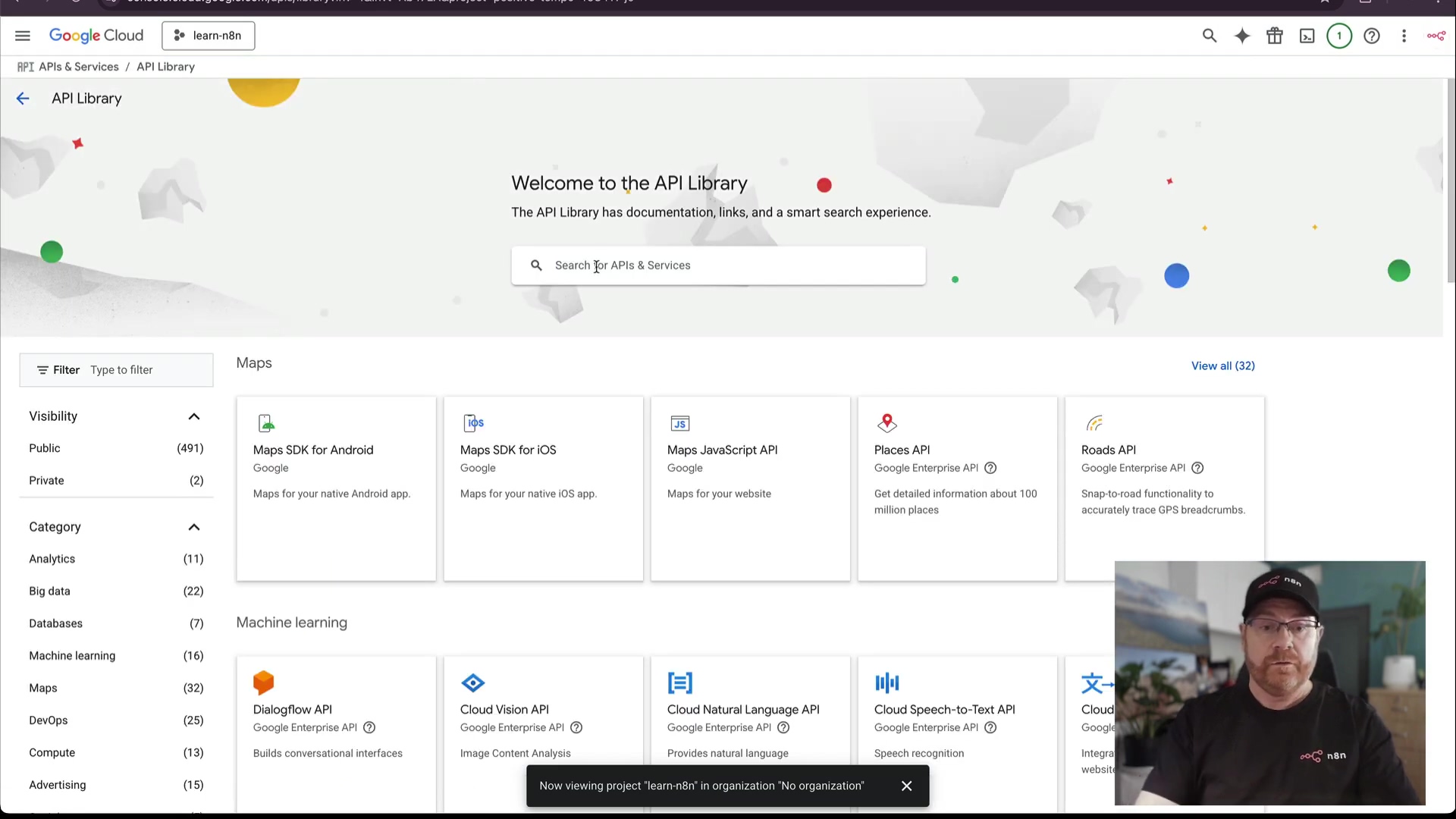 The image shows the Google Cloud 'API Library' interface. A sidebar on the left features 'Filter', 'Visibility' (Public, Private), 'Category' (Analytics, Big data, Databases, DevOps, Gaming, Machine learning, Maps, Compute, Advertising), and 'Product' with various numbers next to each category. The main content area displays 'Welcome to the API Library' with a search bar 'Search APIs & Services'. Below the search bar, several API cards are visible, including 'Maps SDK for Android', 'Maps SDK for iOS', 'Maps JavaScript API', 'Places API', 'Roads API', 'Dialogflow API', 'Cloud Vision API', 'Cloud Natural Language API', 'Cloud Speech-to-Text API', and 'Drive API'. A banner at the bottom states 'Now viewing project "learn-n8n" in organization "No organization"'. A man with glasses and a black cap is visible in the bottom right corner, wearing a black t-shirt with a white logo. The interface is white and gray with blue text.