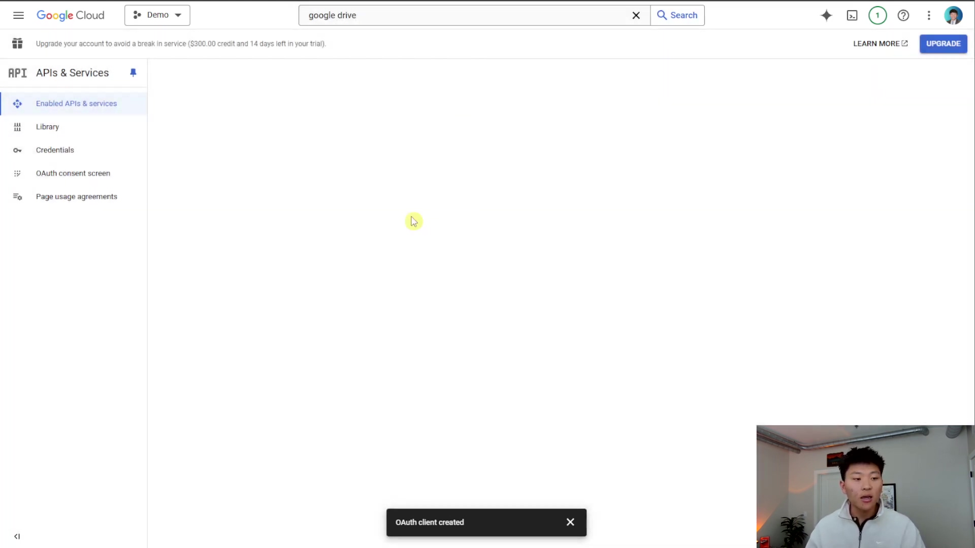 The image displays a Google Cloud console interface with a sidebar menu on the left and a large blank content area on the right. The top bar features the 'Google Cloud' logo, a dropdown labeled 'Demo', a search bar with 'google drive' typed in, and icons for notifications, help, and user account. A banner at the top warns to 'Upgrade your account to avoid a break in service ($300.00 credit and 14 days left in your trial)'. The left sidebar shows navigation items under 'API APIs & Services' including 'Enabled APIs & Services', 'Library', 'Credentials', 'OAuth consent screen', and 'Page usage agreements'. A small black pop-up at the bottom center reads 'OAuth client created'. A person is visible in the bottom right corner, looking at the screen.