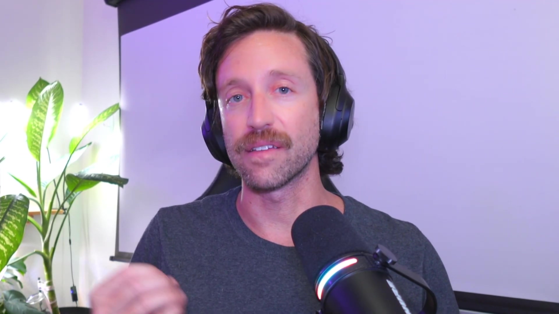 A man with a mustache and short brown hair is shown from the chest up, looking slightly to the right of the camera. He is wearing a dark grey t-shirt and black over-ear headphones. His left hand is partially visible at the bottom left, gesturing with his fingers. Behind him, to the left, is a green plant with large leaves, and to the right, a microphone with a colorful LED light ring is visible. The background is dimly lit with purple and white ambient lighting, suggesting an indoor setting, possibly a home office or studio.