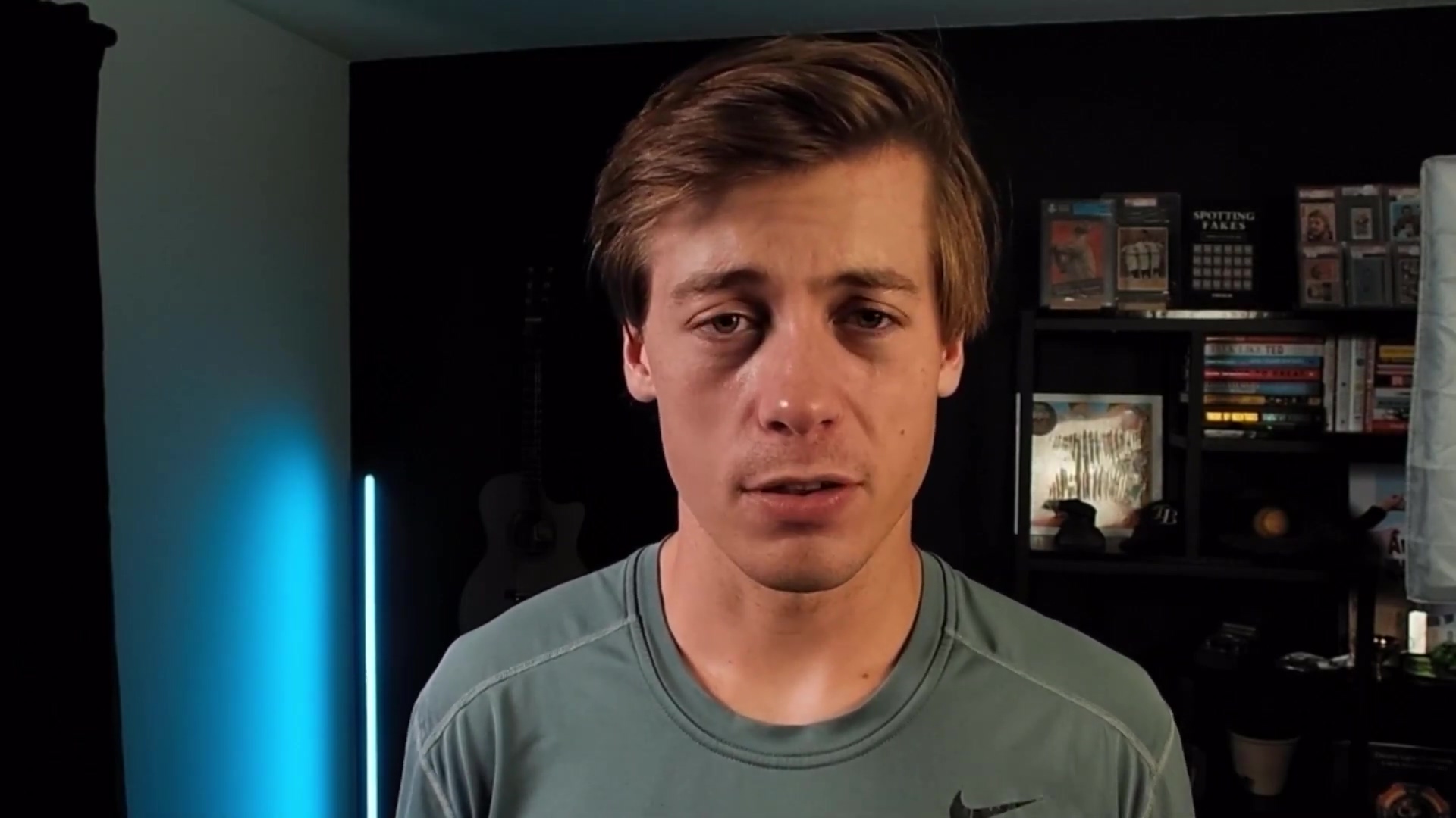 The image shows a young man, likely the video presenter, looking directly at the camera. He has short, light brown hair and is wearing a teal-colored t-shirt with a black Nike logo on the right chest area. His expression is neutral, and he appears to be speaking. The background is dark, with a black wall behind him. To his left, a light-colored acoustic guitar is partially visible. To his right, a black shelf unit holds various items, including what appear to be framed trading cards or collectibles, and some books. A blue light emanates from the left side of the frame, casting a subtle glow.