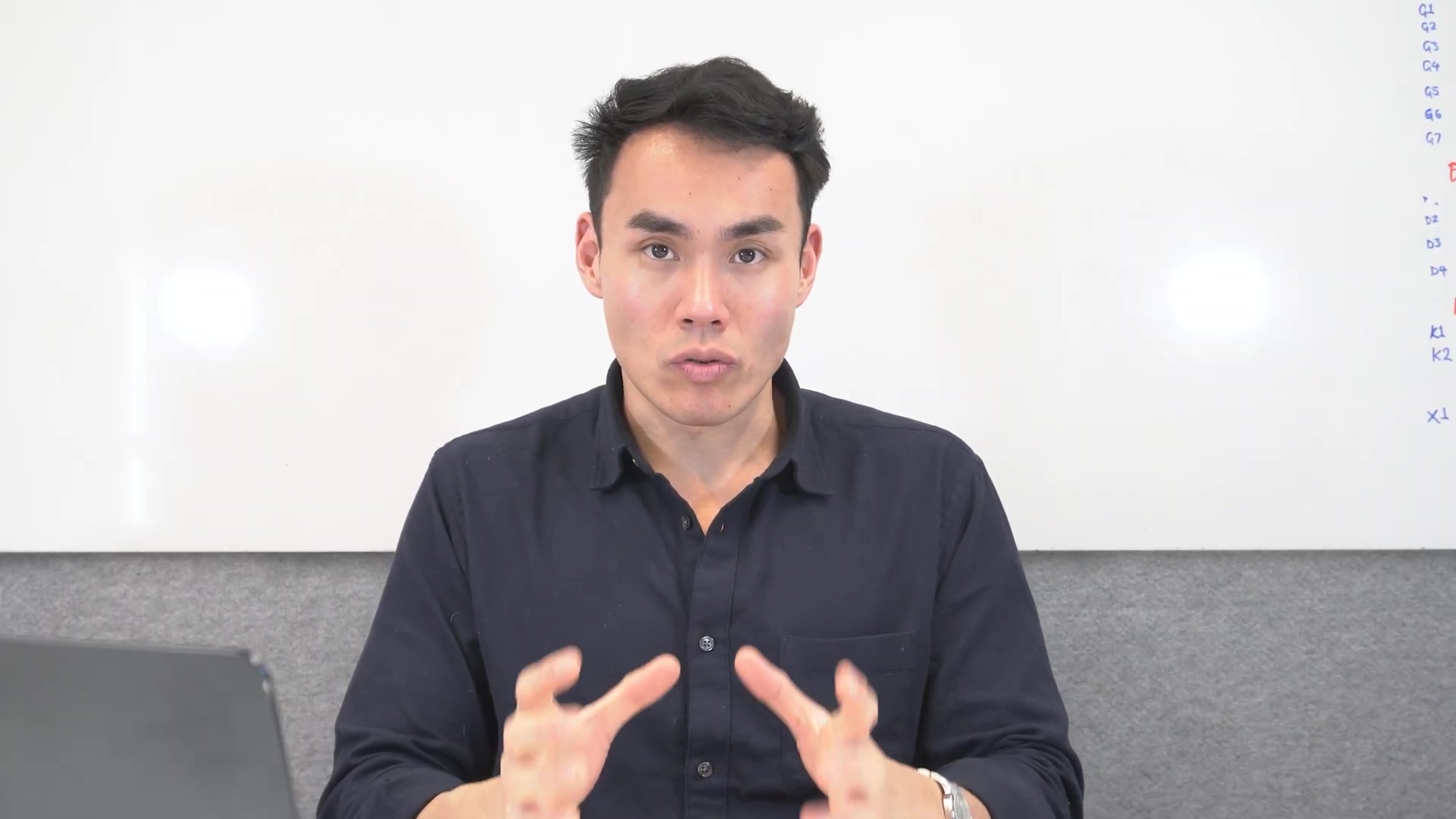A man with short dark hair and a dark blue collared shirt is seated at a desk, looking directly at the camera and speaking. His hands are positioned in front of him, gesturing as he talks. A silver laptop is partially visible on the desk to his left. Behind him is a white board with handwritten notes in blue and red markers, similar to the previous image. The notes are partially visible and include headings like 'KEYCLOAK INTEGRATION', 'DCP', 'DATABASE', and 'DOCS'. The lighting is bright and clear.