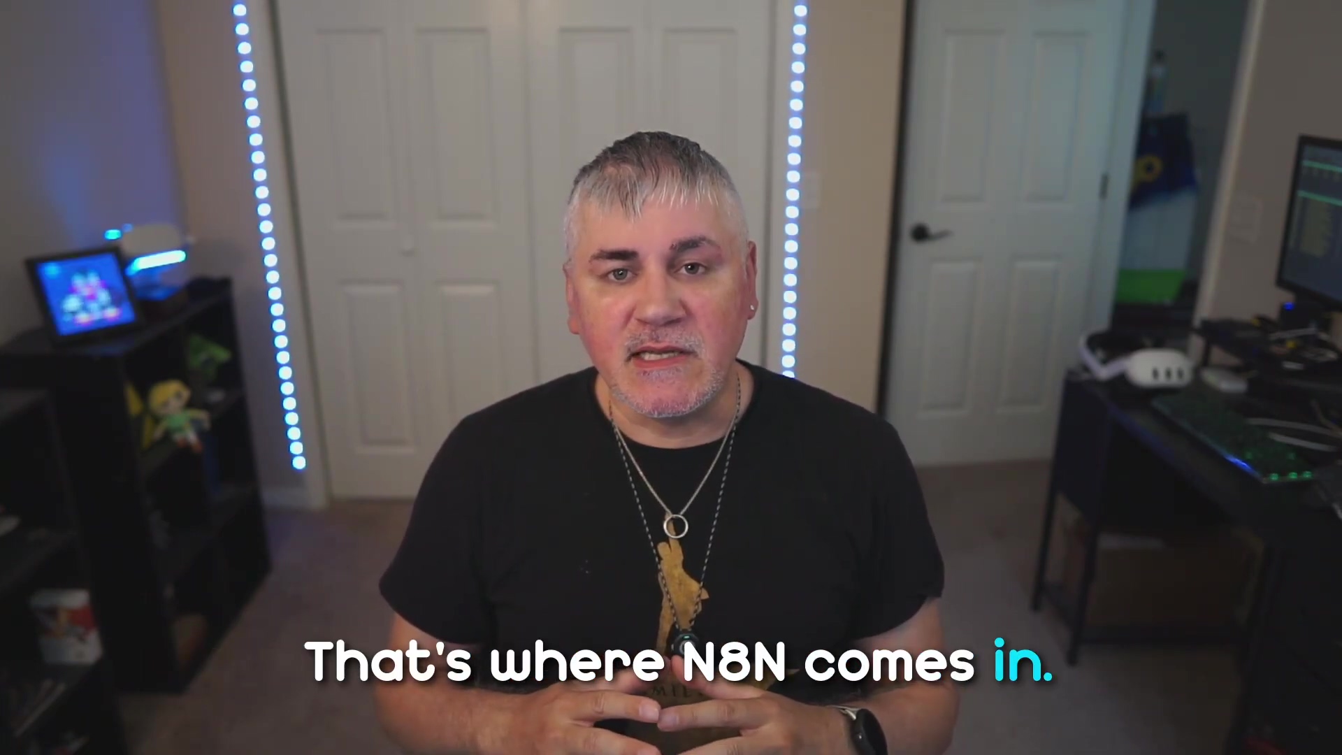 The image shows a man with short, gray and dark hair, wearing a black t-shirt and a necklace, speaking directly to the camera. He is positioned centrally in the frame. Behind him, there are white double doors and a dark shelf unit to his left, which holds various items including what appears to be a blue-lit device and a green-colored object. To his right, a desk with a computer monitor and keyboard is partially visible. The room is lit by blue LED strip lights running vertically on the walls behind him. White text overlays the bottom of the screen, stating "That's where N8N comes in."