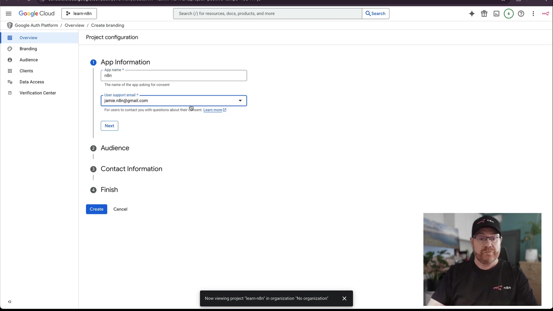The image displays a Google Cloud Platform interface for configuring an OAuth consent screen. The main section, titled "Project configuration," shows "App Information" with fields for "App name" and "User support email." The app name is set to "n8n" and the email is "jamien8n@gmail.com." Below this, there are sections for "Audience," "Contact Information," and "Finish," which are collapsed. On the left sidebar, navigation options include "Overview," "Branding," "Audience," "Clients," "Data Access," and "Verification Center." The top bar shows "Google Cloud" logo, "learn-n8n" project selected, a search bar, and user icons. A small pop-up at the bottom reads "Now viewing project 'learn-n8n' in organization 'No organization'." A man with glasses and a black t-shirt with a logo is visible in the bottom right corner of the screen, looking towards the interface.