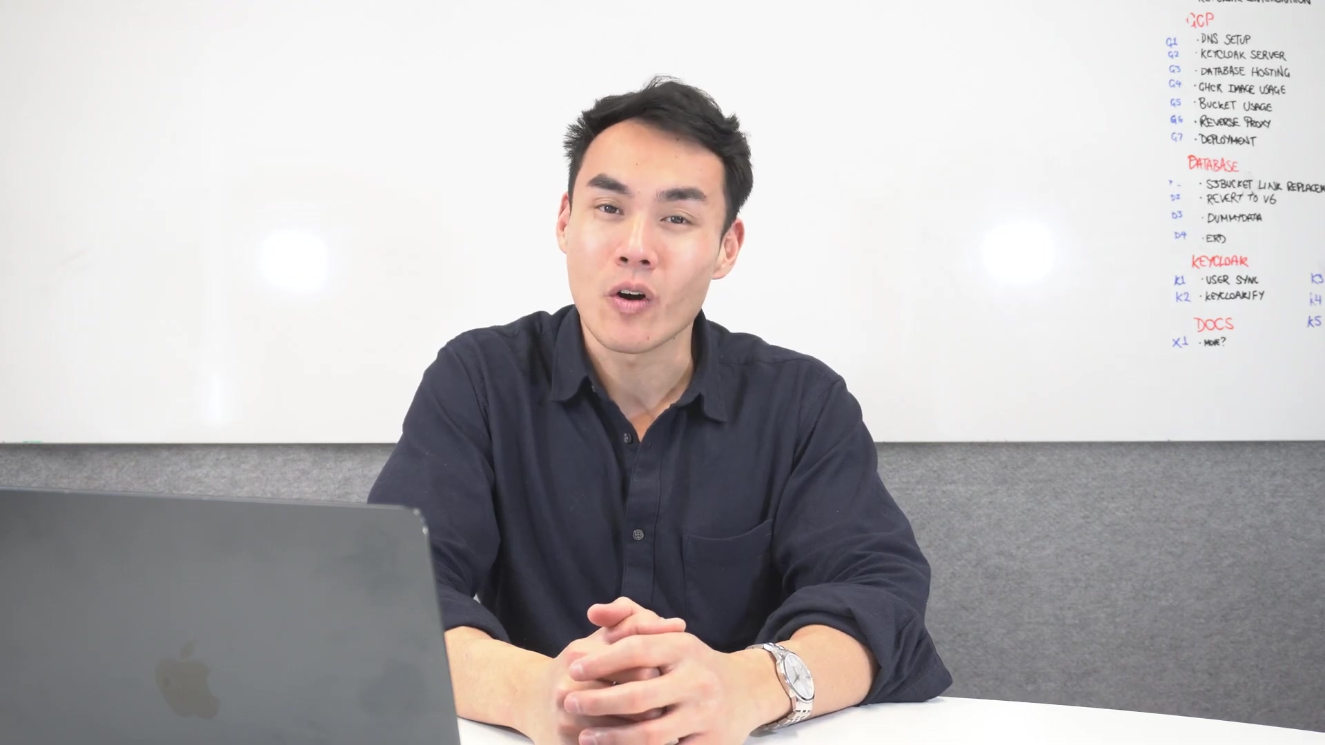 A man with short dark hair and a dark blue collared shirt is seated at a desk, looking directly at the camera and speaking. His hands are clasped together on the desk in front of him. To his left, a silver laptop is partially visible. Behind him is a white whiteboard with handwritten notes in red and blue marker, listing various technical terms and categories such as "XDP," "DATABASE," "KEYCLOAK," and "DOCS." The room appears to be an office setting with a grey wall in the background.