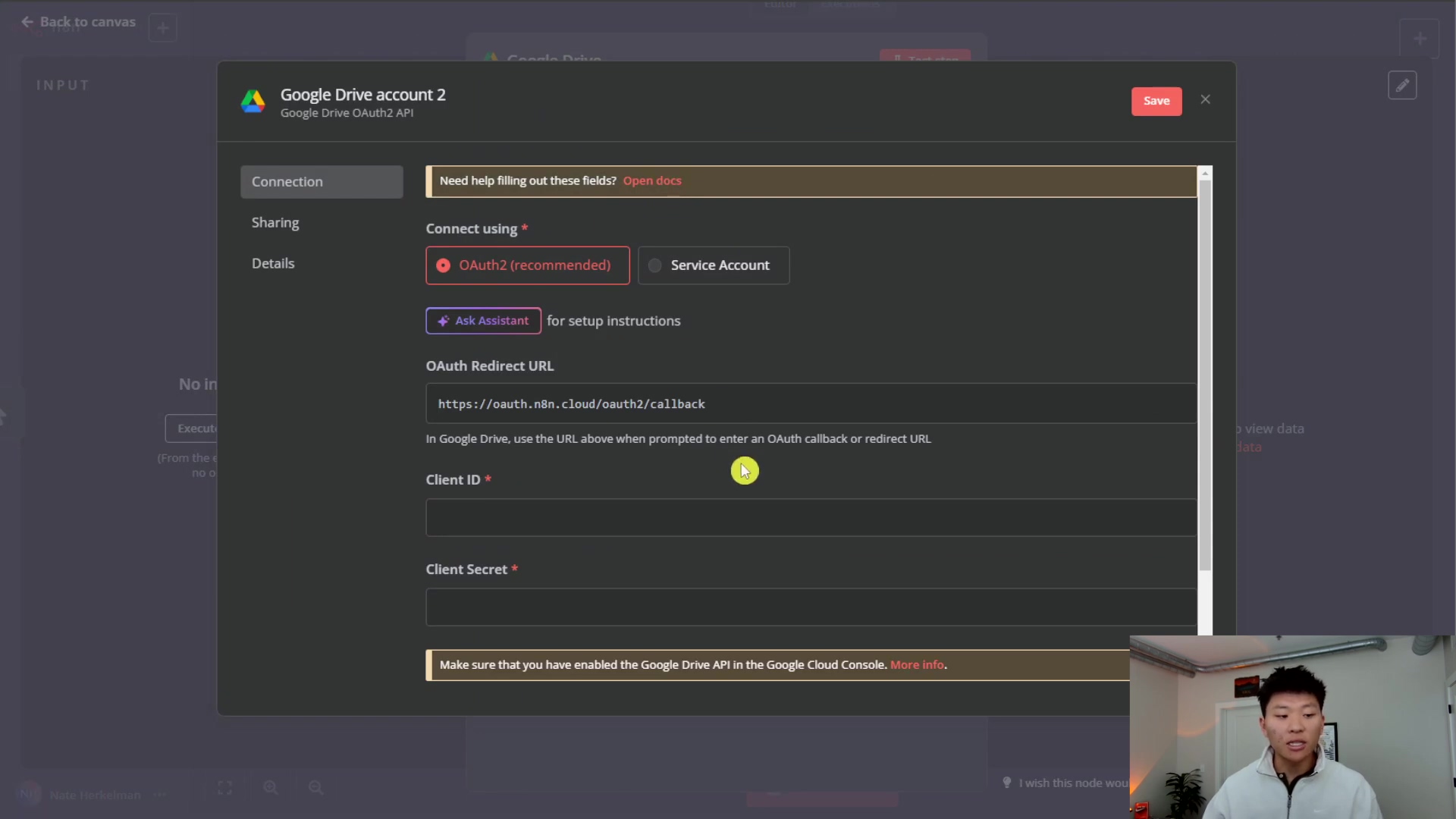 The image shows a modal window within the n8n interface titled 'Google Drive account 2' for configuring Google Drive OAuth2 API credentials. The modal has tabs for 'Connection', 'Sharing', and 'Details'. The 'Connection' tab is active, displaying fields for 'Connect using' with radio buttons for 'OAuth2 (recommended)' and 'Service Account'. 'OAuth2 (recommended)' is selected. Below this, there's a button 'Ask Assistant' for setup instructions. A crucial field, 'OAuth Redirect URL', is pre-filled with 'https://oauth.n8n.cloud/callback'. Instructions state: 'In Google Drive, use the URL above when prompted to enter an OAuth callback or redirect URL.' Further down, there are empty input fields for 'Client ID *' and 'Client Secret *', both marked as required. A note at the bottom advises: 'Make sure that you have enabled the Google Drive API in the Google Cloud Console. More info.' A person is visible in the bottom right corner, looking at the screen.