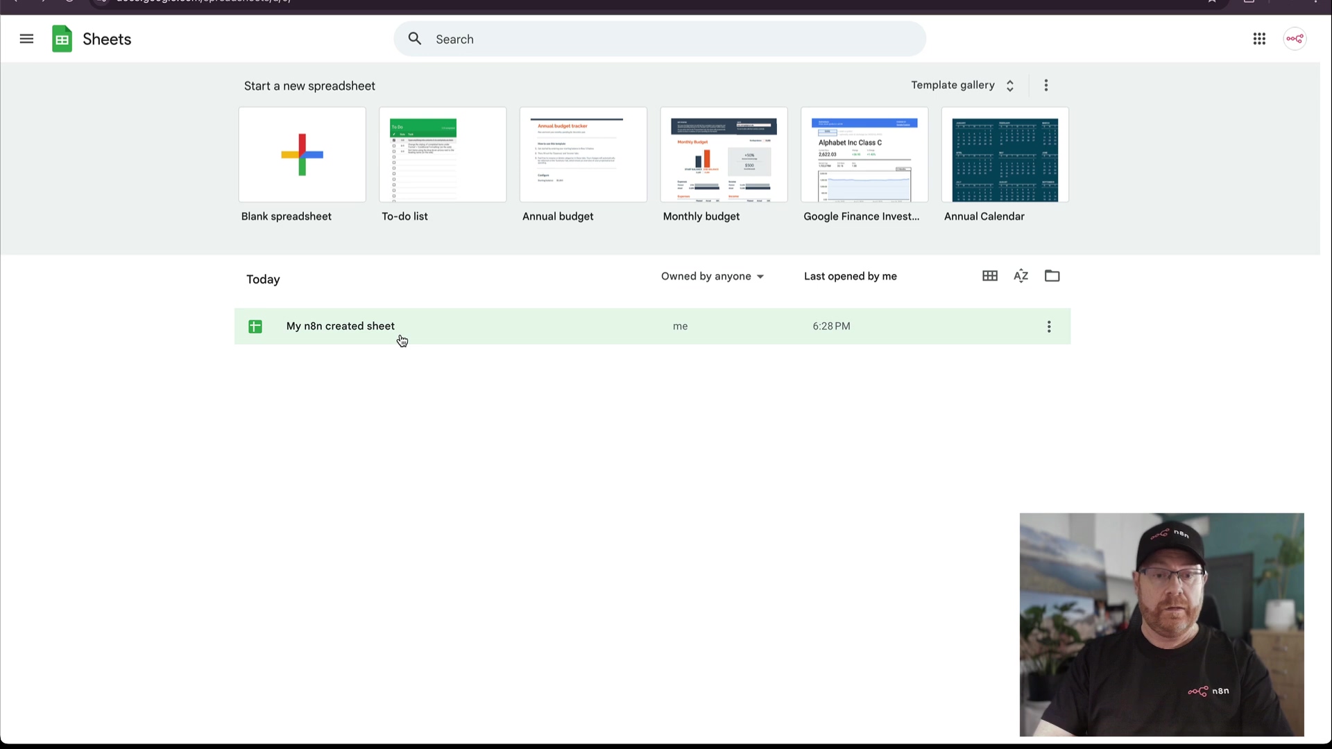 The image displays the Google Sheets homepage, showing various spreadsheet templates and recently opened files. At the top, a search bar is visible. Below it, options like 'Start a new spreadsheet' with a large plus icon for 'Blank spreadsheet', followed by 'To-do list', 'Annual budget', 'Monthly budget', 'Google Finance Investment Tracker', and 'Annual Calendar' templates. Under 'Today', a file named 'My n8n created sheet' is listed, showing 'me' as the owner and '6:29 PM' as the last opened time. The bottom right corner of the screen shows a man with glasses and a beard, wearing a black t-shirt with a white logo, looking towards the screen. His presence indicates a demonstration or tutorial context.