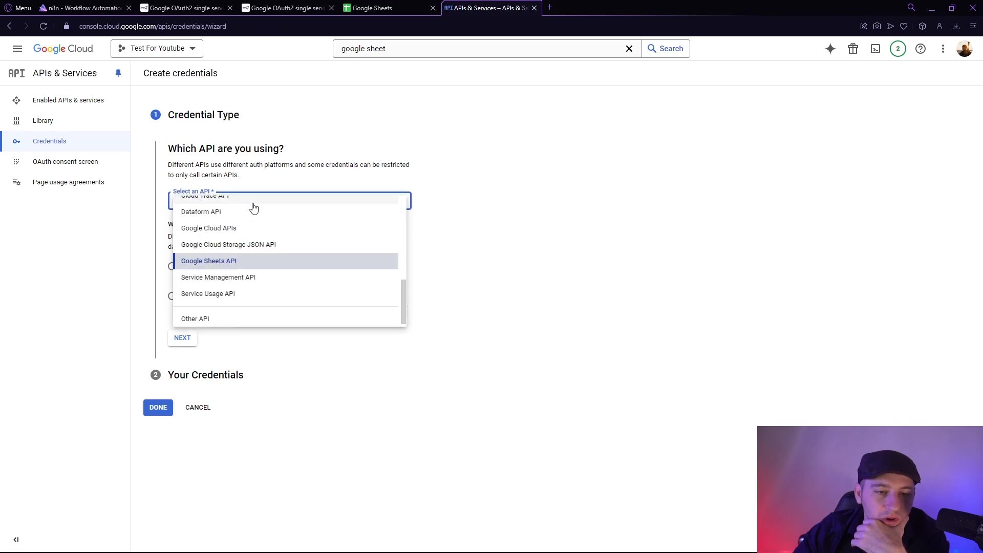The image displays the Google Cloud Console interface, specifically the 'Create credentials' section. The main content area shows a dropdown menu labeled 'Select an API' with 'Google Sheets API' highlighted and selected. Below this, there's a 'NEXT' button. On the left sidebar, 'Credentials' and 'OAuth consent screen' are visible. A person is visible in the bottom right corner, looking at the screen.