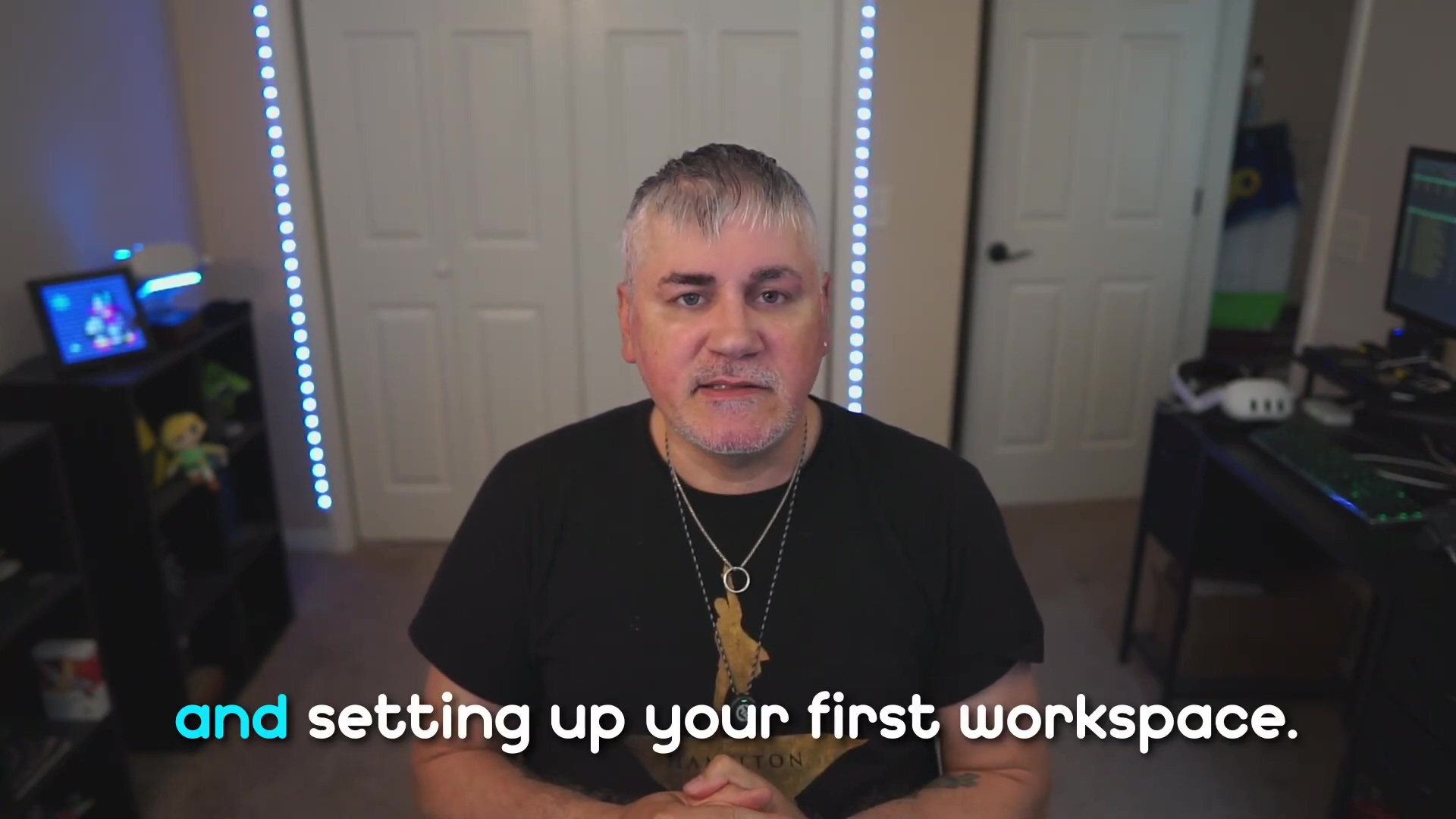 The image displays the same man from the previous frame, with short, graying hair and a beard, wearing a black t-shirt with a graphic. He is looking directly at the camera with a slight smile. His hands are clasped together in front of him. The background is consistent with the previous image, featuring white walls, a closed white door on the right, a dark shelf unit with various items on the left, and vertical blue LED strip lights on both sides. A bright blue subtitle at the bottom reads "and setting up your first workspace."