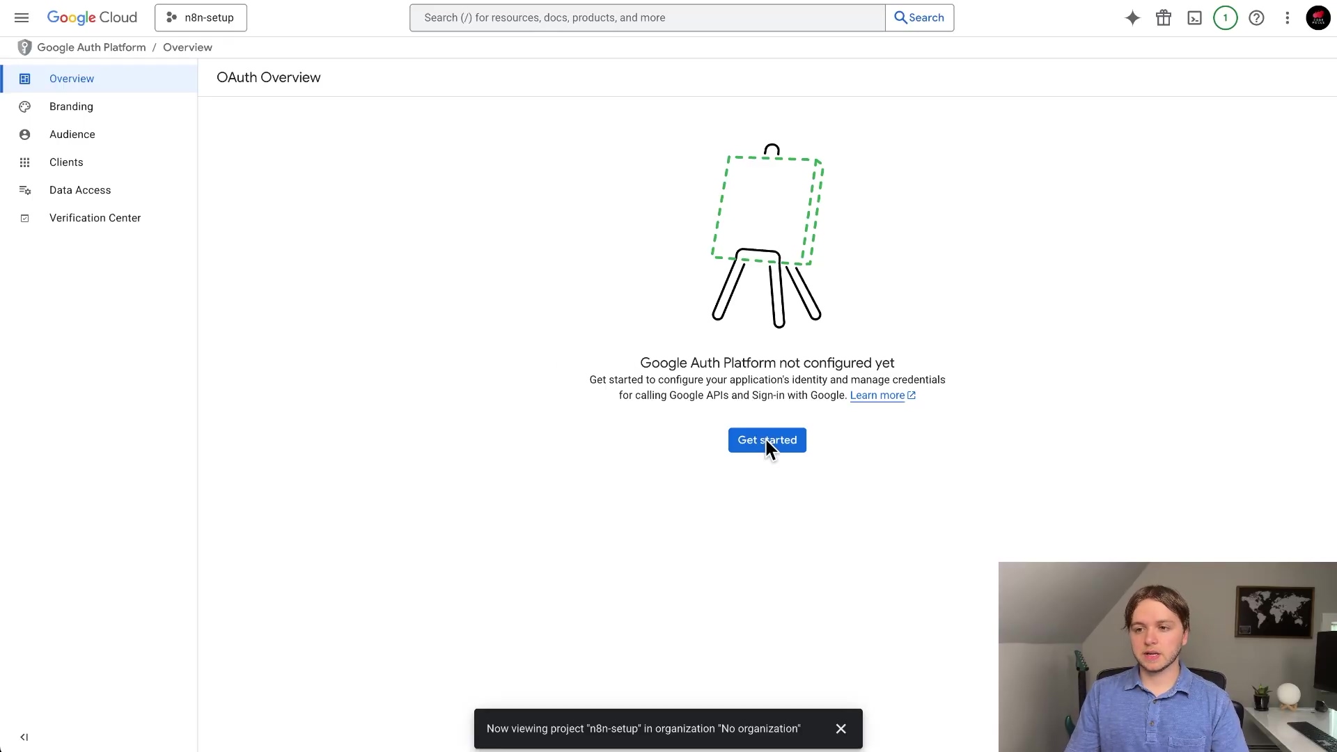 The image displays the Google Cloud Platform interface, specifically the 'OAuth Overview' section. A large, stylized icon of an easel with a dashed outline is centered on the screen, indicating that the Google Auth Platform is not yet configured. Below the icon, there's a message prompting the user to get started, with a prominent blue 'Get started' button. The left sidebar shows navigation options like 'Overview', 'Branding', 'Audience', 'Clients', 'Data Access', and 'Verification Center'. At the top, there's a search bar and various Google Cloud branding elements. A person is visible in the bottom right corner of the screen, looking at the interface.