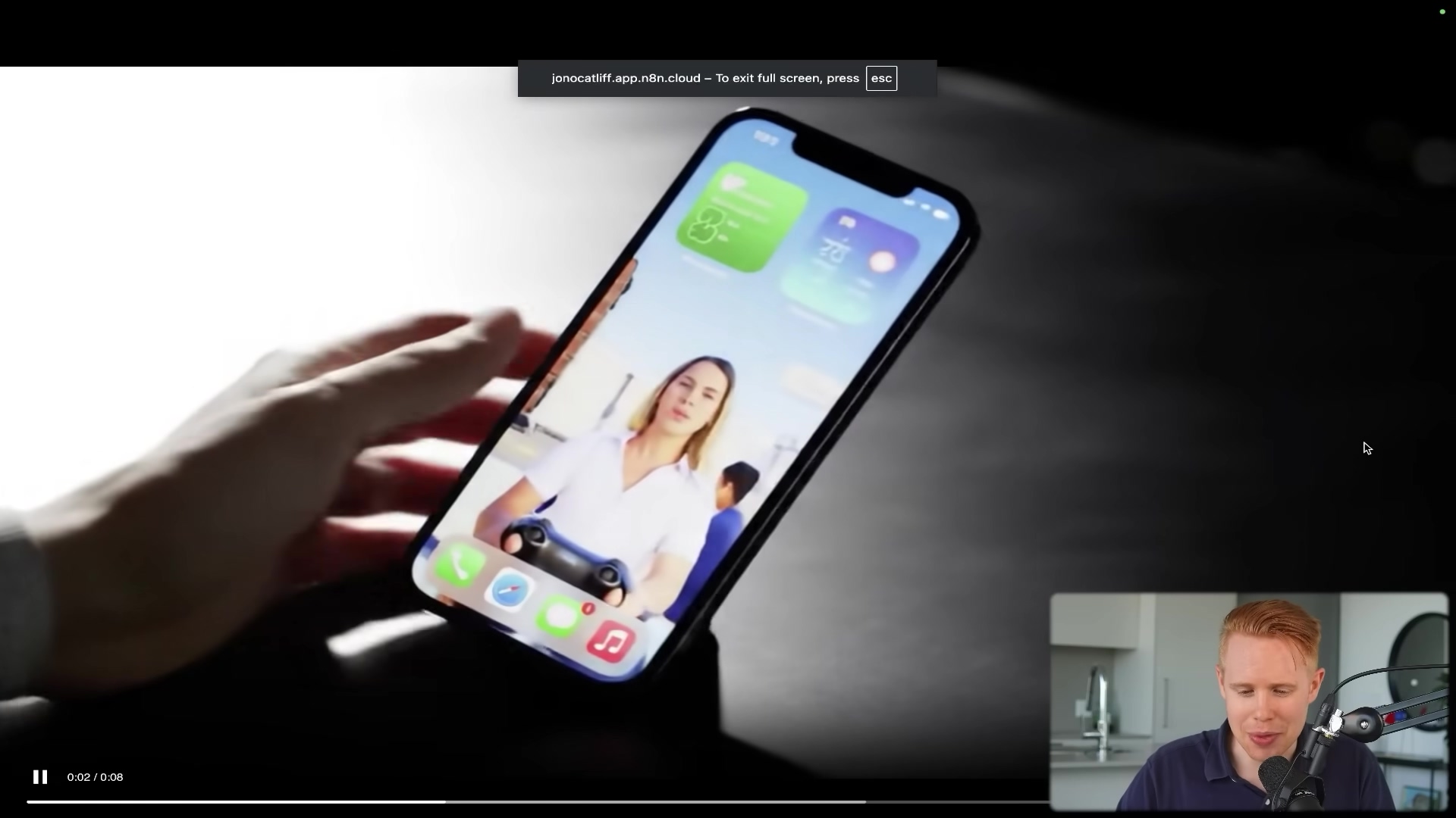 The image shows a close-up of a smartphone (likely an iPhone) held by a hand, displaying its screen. The screen shows a woman's face and a video game controller, suggesting video content. The phone's interface includes app icons and widgets, with a weather widget showing '70°' and a calendar widget. A banner at the top of the screen reads 'jonocatliff.app.n8n.cloud – To exit full screen, press esc'. A man with short red hair is visible in the bottom right corner of the overall frame, looking at the screen with a surprised expression. A video player progress bar is visible at the bottom of the screen, indicating '0:02 / 0:08'.