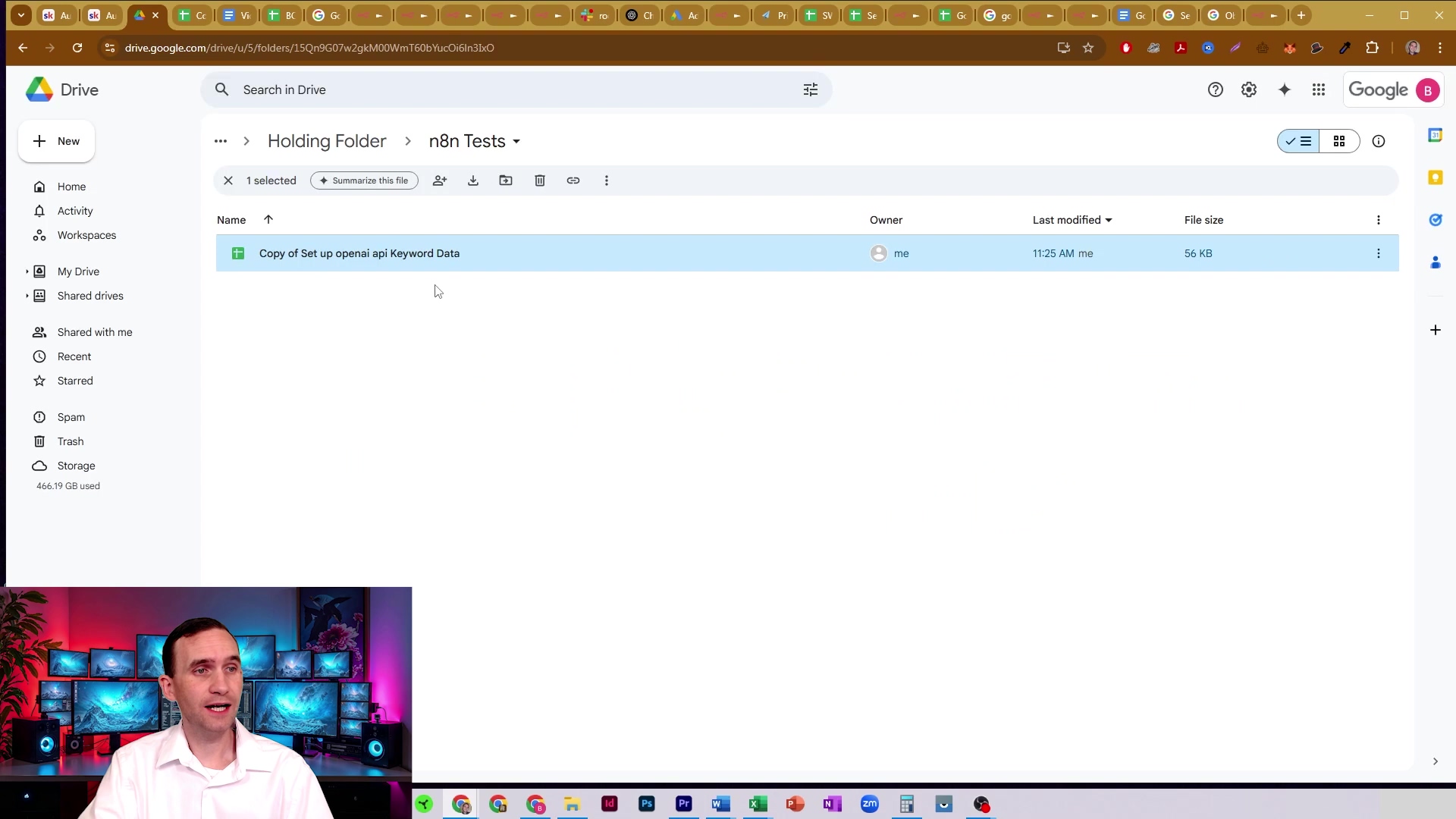The image shows a Google Drive interface displaying a folder named "n8n Tests" within "Holding Folder." A single Google Sheet file, "Copy of Set up openai api Keyword Data," is selected and highlighted in blue. Details about the file, such as "Owner: me," "Last modified: 11:25 AM," and "File size: 56 KB," are visible. The left sidebar shows Google Drive navigation options like "My Drive," "Shared with me," "Recent," "Starred," and "Storage." A man is visible in the bottom left corner, speaking, with multiple computer monitors behind him.