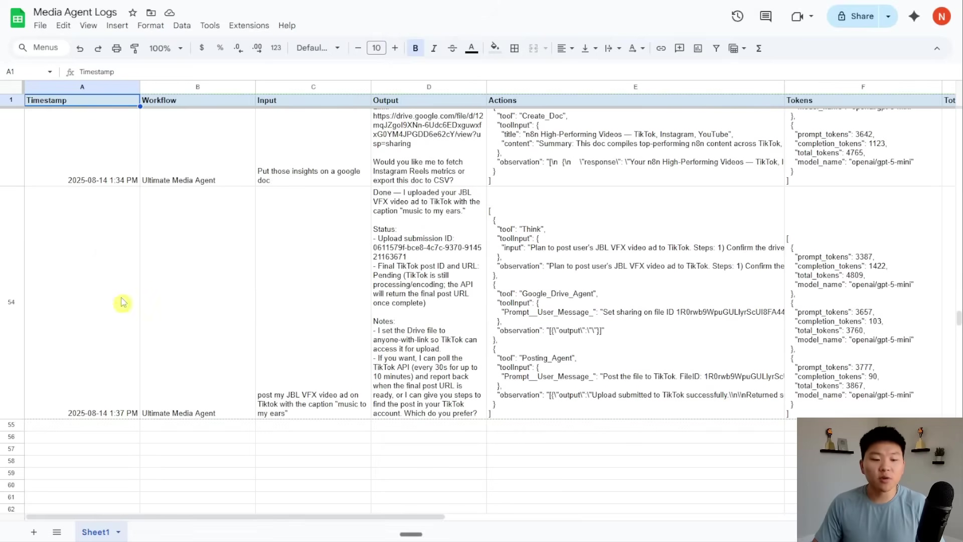 The image displays a Google Sheet titled "Media Agent Logs" with columns for Timestamp, Workflow, Input, Output, Actions, and Tokens. The sheet shows entries for August 14, 2024, with details of an "Ultimate Media Agent" workflow. The 'Input' column contains text like "Put those insights on a Google doc" and "post my JBL VFX video ad on TikTok with the caption 'music to my ears'". The 'Output' column shows responses and status updates, including file processing, sharing settings, and a question about naming a file. The 'Actions' column details the steps taken by the agent, such as "Create Doc", "Post to TikTok", and "Google Drive Agent". The 'Tokens' column provides data on prompt_tokens, completion_tokens, total_tokens, and model_name (e.g., "openai/gpt-3.5-mini"). The bottom right of the image shows a man with short dark hair and a black t-shirt, looking at the screen and talking. A microphone is visible in front of him.